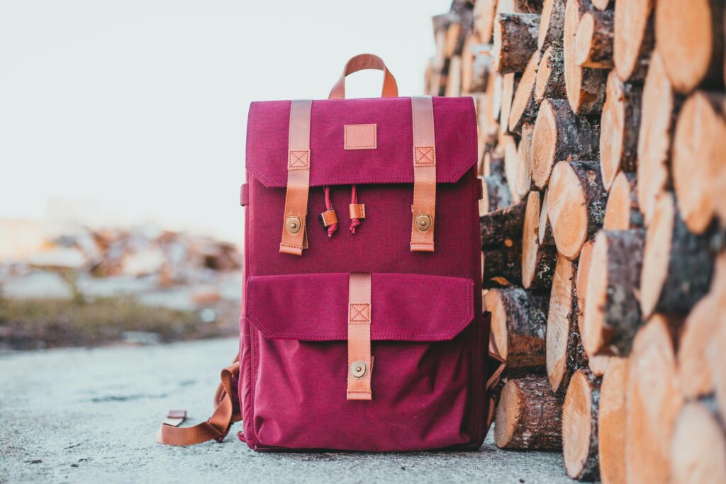 Pink backpack with leather straps next to stacked firewood, showcasing outdoor adventure gear.